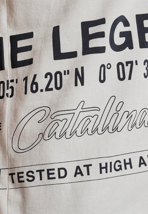 Bela tkanina z drznim črnim napisom. Vključuje koordinate, "Catalina" in "TESTIRANO NA VISOKIH VIŠINAH." Gladka tekstura, sproščen dizajn.