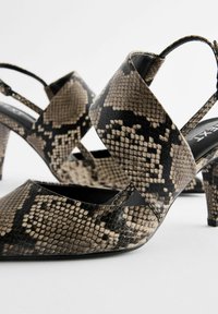 Des chaussures à talons hauts noires et beige avec un motif serpent, à bout pointu et à brides croisées à la cheville, fabriquées en matériau synthétique texturé.