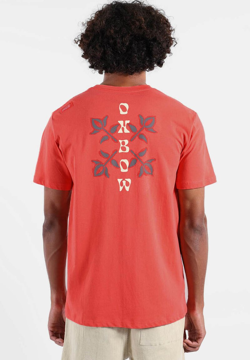 T-shirt en coton rouge avec un motif floral et le mot "OKBOW" au centre. Manches courtes et coupe décontractée.