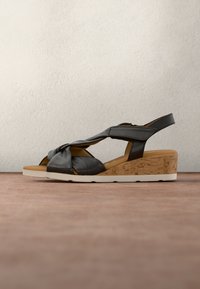 Sandale compensée en cuir noir avec brides torsadées, talon texturé liège, semelle intérieure marron et semelle extérieure en caoutchouc blanc sur une surface marron.