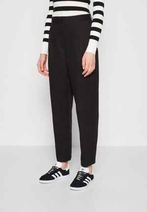 Persoon draagt zwarte taps toelopende broek, zwart-wit gestreepte top met lange mouwen en zwarte Adidas sneakers met witte strepen en zolen.