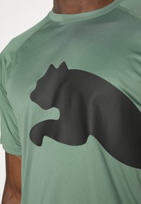 Zielona sportowa koszulka z matowym nadrukiem w kształcie czarnego kota z przodu. Materiał jest gładki i ma okrągły dekolt.