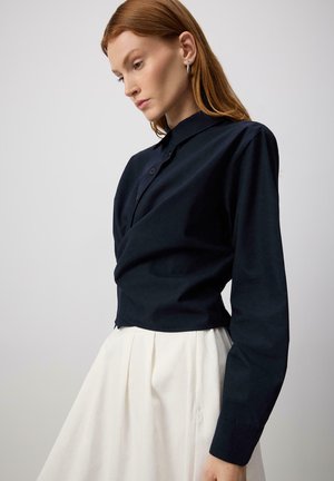 Žena s červenými vlasmi, ktorá má na sebe strieborné kruhové náušnice, čiernu košeľu s dlhými rukávmi na gombíky a bielu plisovanú sukňu na jednoduchom pozadí.