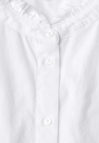Camisa blanca con un cuello con volantes, que presenta una fila de pequeños botones transparentes y una tela texturizada con sutiles rayas diagonales.