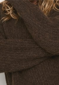 Brun ulltröja med en texturerad, fluffig yta, vida ärmar och en lös passform. Närbilden betonar tygets mjukhet.