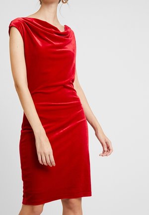 Ärmelloses rotes Samtkleid mit drapiertem Ausschnitt, tailliert und knielang an einer stehenden Person vor einem einfarbigen hellen Hintergrund.
