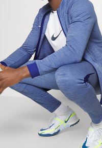 Blauwe sportieve tracksuit van zacht materiaal, met een aansluitend ontwerp en marineblauwe accenten. Schoenen zijn wit met blauwe en neon gele details.