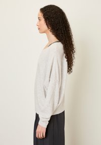 Svetlo sivi oversized pulover z mehko teksturo, širokim ovratnikom in spuščenimi rameni. Nošen preko temno sive majice brez roka, v kombinaciji z dolgimi krili.
