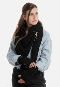 Jonge vrouw in een lichtdenim jas, zwarte gebreide sjaal vastgezet met een houten sluiting, en bijpassende zwarte vingerschoenen, die naar rechts kijkt.