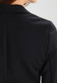 Musta räätälöity bleiseri, jossa on jämäkkä muotoilu, sileä pinta ja terävät läppäkoristeet, esittäen puhtaita linjoja ja klassista siluettia.