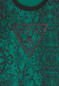 Camiseta con estampado de serpiente en verde y negro, con un logo triangular "GUESS?" en negro brillante, y cuello redondo de canalé negro.