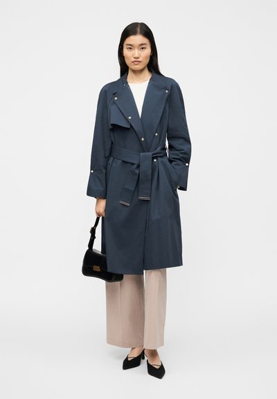 Μπλε ναυτικό παλτό με ζώνη στη μέση, μεγάλο γιακά και κουμπιά με πατιλέτα. Συνδυάζεται με ανοιχτό μπεζ παντελόνι φαρδιάς γραμμής και μαύρη τσάντα.