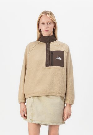 Beige fleece genser med høy krage i brun, kvart glidelås og venstre brystlomme med hvit logo. Strukturen er myk og behagelig.