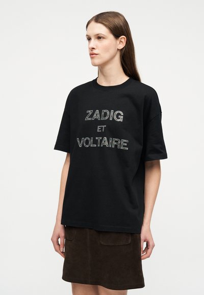 Νεαρή γυναίκα με ίσια καστανά μαλλιά, που φοράει μια μαύρη υπερμεγέθη κοντομάνικη μπλούζα με το κείμενο "ZADIG ET VOLTAIRE" και μια σκούρα καφέ φούστα.