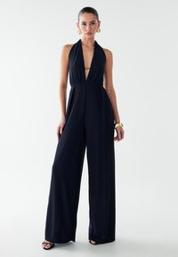 Mono de marina con cuello halter, escote en V profundo, piernas anchas y tela texturada. Incluye una pulsera dorada y grandes pendientes redondos.