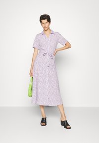 Vestido camisola lilás claro com padrão floral, mangas curtas, frente com botões e cintura atada. Combinado com mules pretas de ponta aberta e uma bolsa verde.
