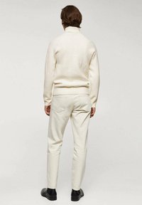 Weißer Strickpullover mit hohem Kragen, kombiniert mit beigen Slim-Fit-Hosen und schwarzen Stiefeletten. Schlichtes Design mit strukturiertem Stoff.
