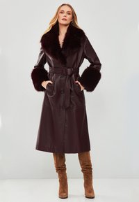 Cappotto in ecopelle bordeaux con ampio colletto e polsini in pelliccia, vita accentuata da cintura, chiusura a bottoni e design lungo fino al ginocchio.