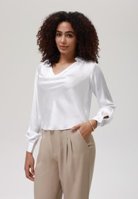 Blusa de satén blanco con escote drapeado y mangas largas, combinada con pantalones de tela beige con pliegues. Textura suave y diseño limpio.