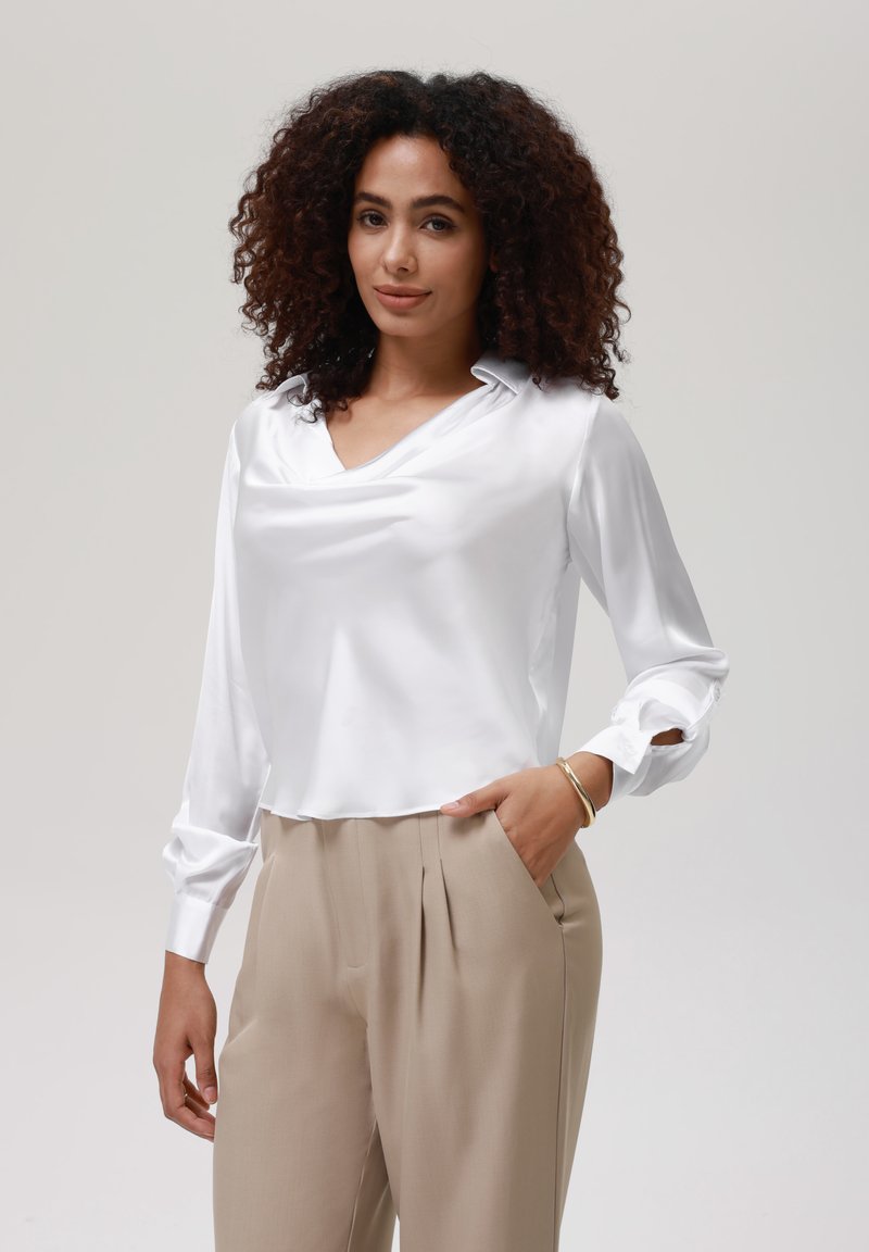 Blusa de satén blanco con escote drapeado y mangas largas, combinada con pantalones de tela beige con pliegues. Textura suave y diseño limpio.