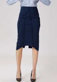 Jupe crayon bleu marine en tissu extensible, avec des coutures angulaires et un ourlet asymétrique. Portée avec des escarpins noirs pointus.