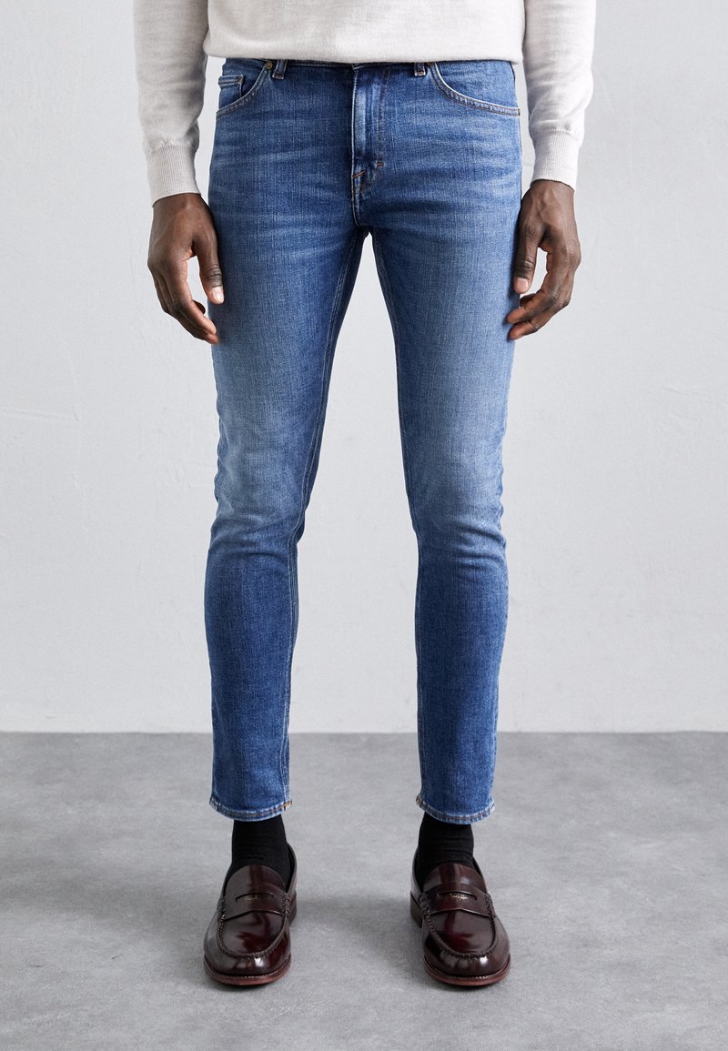 Slim-fit modré džíny s vybledlým vzhledem, pět kapes a zipový zip. Kombinované s tmavými mokasíny a černými ponožkami.