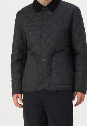 Mężczyzna w czarnej pikowanej kurtce z czarnym kołnierzem, zapięciem na guziki, kieszeniami oraz w czarnych spodniach na białym tle.