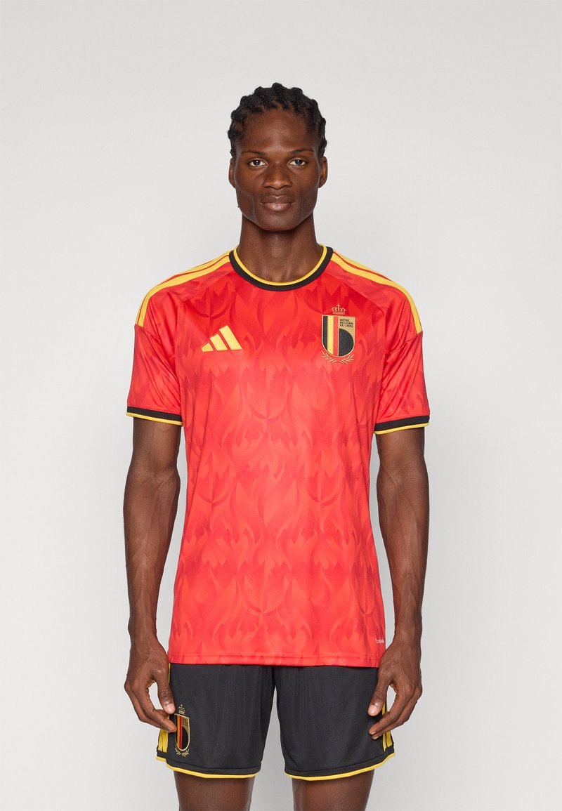 Mannelijke atleet draagt een rood Belgisch voetbalshirt met vlammenpatroon, zwarte korte broek, staand tegen een effen grijze achtergrond.