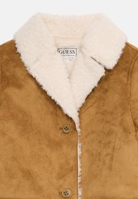 Chaqueta de ante marrón con cuello y forro de sherpa crema, dos botones marrones y etiqueta de la marca Guess visible en el interior del cuello.