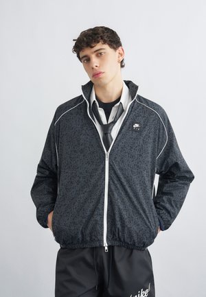 Mladý muž s kučeravými vlasmi nosí čiernu vzorovanú bundu Nike Air, biele tričko, čiernu kravatu a čierne nohavice Nike, stojaci s rukami v vreckách.