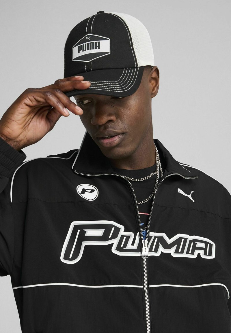 Crna sportska jakna s bijelim detaljima i velikim "PUMA" logom, u kombinaciji s crnom i bijelom kapom koja prikazuje PUMA logo.
