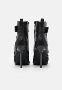 Botines negros de tacón alto, hechos de cuero suave, con un diseño curvado, cremalleras laterales y un acento de correa cerca de la parte superior. Tacón de aguja visible.