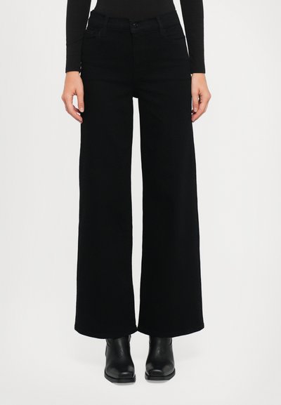 Jean noir à jambe large en denim, avec poches avant, fermeture à bouton et finition soignée. Porté avec des bottines noires.