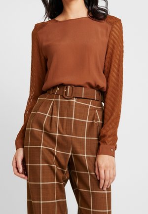 Vrouw draagt een bruine blouse met gestructureerde kanten mouwen, gecombineerd met hoge taille bruin geruite broek en bijpassende riem.