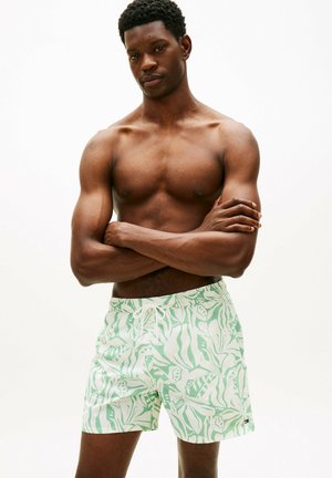 Uomo senza maglietta con le braccia incrociate che indossa pantaloncini da bagno color verde chiaro e bianco con motivo, su sfondo bianco uniforme.