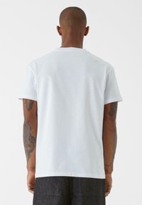 T-shirt bianco in cotone con collo rotondo, maniche corte e vestibilità rilassata. Presenta una textura liscia e un design minimalista.