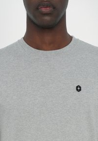 Tricou gri din bumbac cu guler rotund, având un mic patch hexagonal negru cu un logo alb în zona stângă a pieptului. Textură netedă, croială casual.