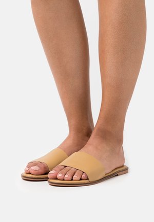 Sandales à enfiler en cuir beige avec une large bride supérieure, design à bout ouvert, semelle plate, et détails minimalistes, portées sur des pieds nus.