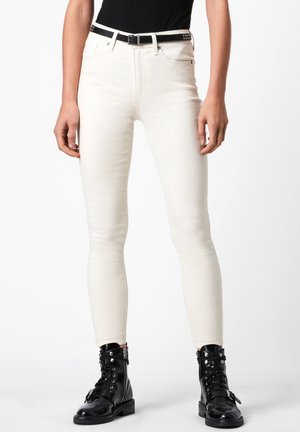 Białe jeansy o dopasowanym kroju połączone z czarnym paskiem z ćwiekami oraz czarnymi skórzanymi sznurowanymi wojskowymi butami na stojącej osobie.