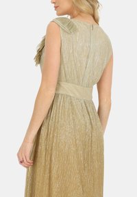 Krótka złota sukienka z teksturowanej tkaniny, z falbaną na ramieniu, dopasowaną talią i luźnym dołem do połowy łydki. Widoczny zamek błyskawiczny z tyłu.