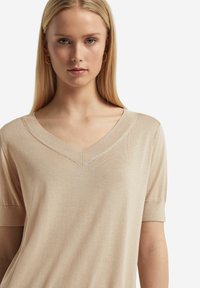 Beżowy sweter typu v-neck z krótkimi rękawami. Gładki, lekki materiał ma subtelny połysk i minimalistyczny design. Brak widocznych wzorów lub akcentów.