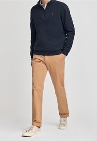 Námořnický svetr na zip s ribovanými manžetami, spárovaný s tan kalhotami. Boty jsou v světlé béžové barvě. Svetr má na hrudi malé červené logo.