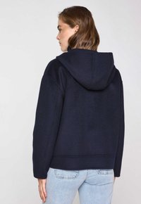 Felpa con cappuccio blu navy, dal tessuto morbido, con taglio oversize e orlo a coste, indossata su jeans azzurri chiari. Vista dalla schiena con un design semplice.