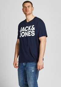Navyblaue Baumwoll-T-Shirt mit "JACK & JONES" in fetter, weißer Schrift. Lockere Passform, kurze Ärmel, Rundhalsausschnitt, kombiniert mit ausgebeulten Jeans.
