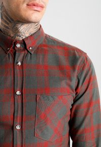Czerwona i szara koszula flanelowa w kratę z długimi rękawami, z kołnierzykiem zapinanym na guziki oraz jedną kieszenią na piersi. Teksturowany materiał o miękkim wykończeniu.