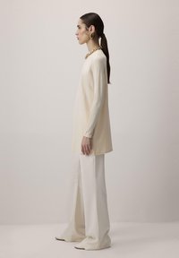 Krém színű, hosszú ujjú felső, széles fehér nadrágokkal párosítva. A felső sima textúrájú és kerek nyakú; figyelemre méltó arany lánc nyaklánc egészíti ki.