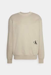 Sweatshirt beige clair avec un col rond, des poignets et un ourlet côtelés. Présente un logo noir vertical et un design de texte sur le devant. Texture de tissu douce.