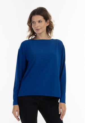 Maglia blu a maniche lunghe con scollatura ampia, morbida al tatto e vestibilità rilassata. Abbinata a pantaloni neri, con cuciture minime e senza motivi visibili.