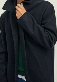 Cappotto di lana scuro con colletto alto, chiusura frontale con bottoni e tasche laterali. Indossato sopra un maglione verde, caratterizzato da una texture morbida.