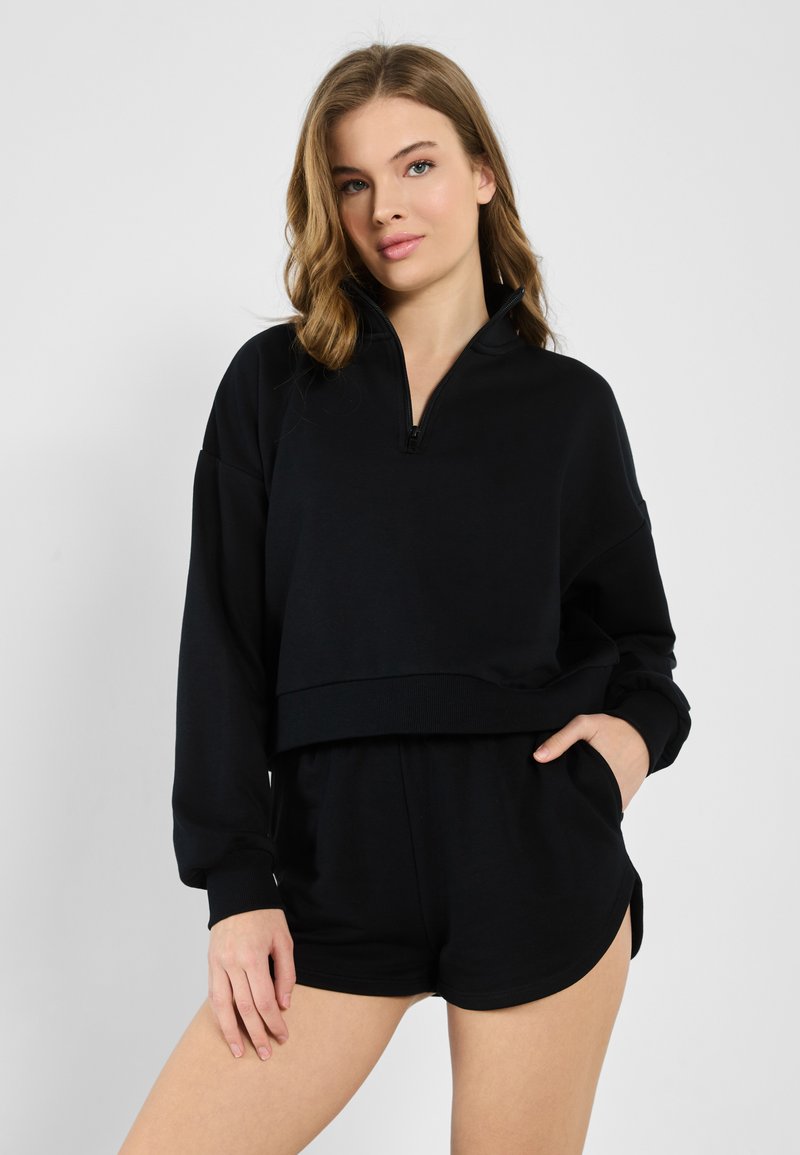 Svart zip-up sweatshirt med en kortad design, långa ärmar och ribbade muddar, kombinerad med svarta shorts. Mjukt tyg med en slät textur.